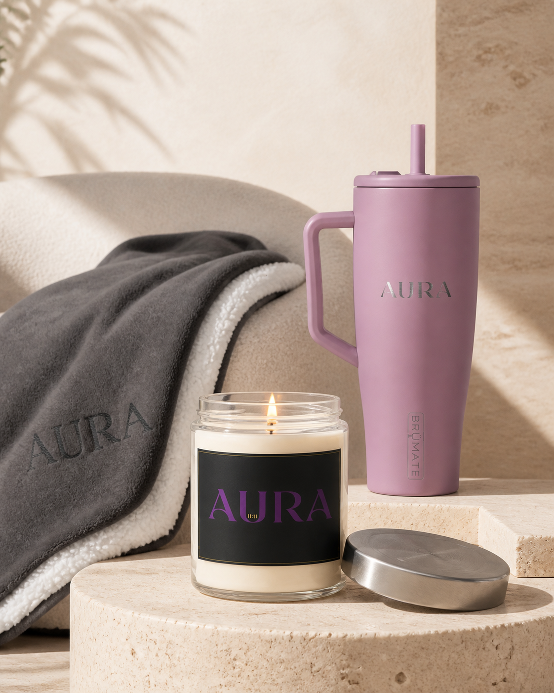 AURA candle with black and violet label, lilac AURA tumbler and embossed AURA blanket on travertine